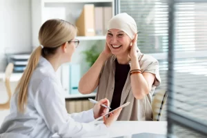 Mulher branca de lenço na cabeça e sorrindo sentada em frente a uma médica branca, ressaltando a importância do diagnóstico precoce do câncer.